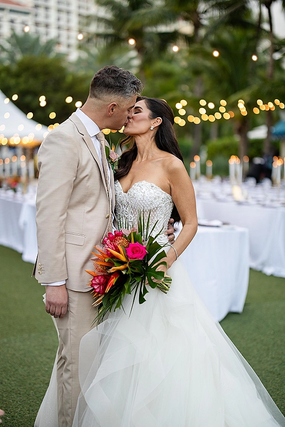 Wedding kiss portrait of bride in strapless lace gown and groom in beige suit, holding tropical bouquet under string lights and palms
