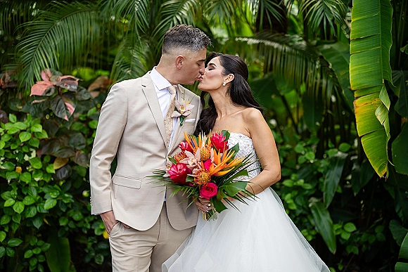 Wedding kiss portrait of bride and groom kissing, bride holding bouquet with veil and strapless dress, framed by tropical greenery and palms