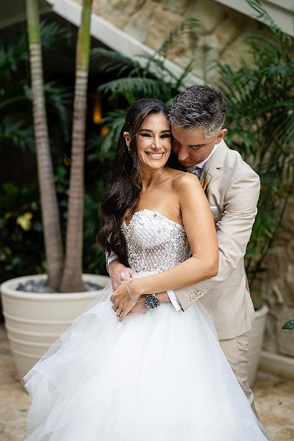 Couple portrait of bride and groom embrace, her strapless beaded ball gown glowing as he hugs her in a tropical courtyard with palm trees