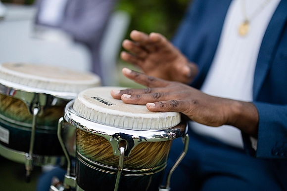 Wedding drummer playing bongo drums in a blue suit with necklace, hands mid-beat, with outdoor greenery and blurred guests behind