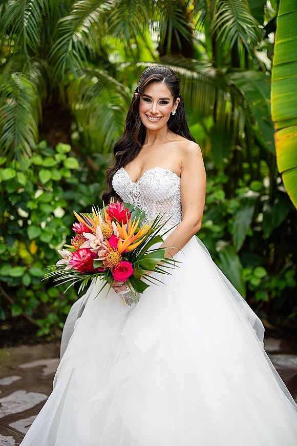 Bridal portrait of a smiling bride holding a tropical bridal bouquet, wearing a strapless beaded tulle gown amid palm leaves on a garden path