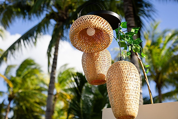 Wedding lighting decor with rattan pendant lights and hanging bulbs on a metal fixture with greenery vine against palm trees and blue sky