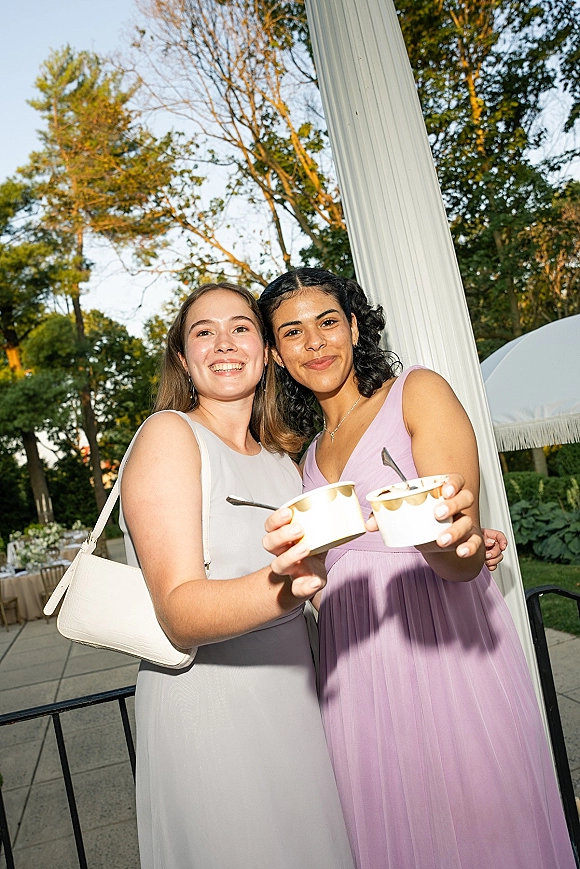 Wedding guest photo of two women in lavender and light gray dresses holding dessert cups on a garden patio with white umbrellas