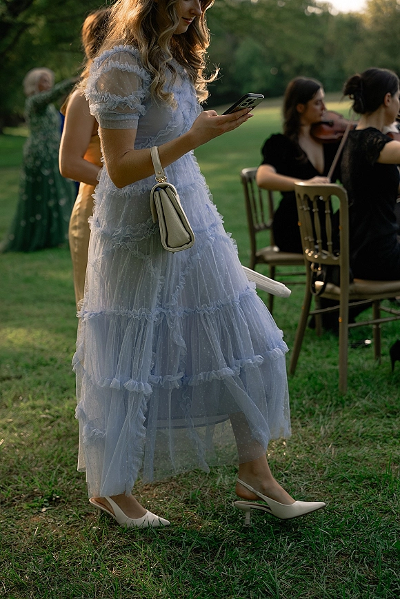 Wedding guest outfit in a blue wedding guest dress with puff sleeves and polka dot tulle, holding a phone beside wooden chairs and violin on a lawn