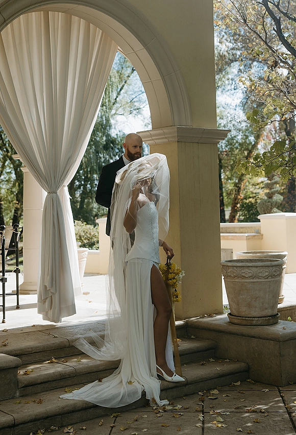 Couple portrait of bride and groom under a stone archway, bride in corset wedding dress with veil and yellow bouquet, groom in black suit