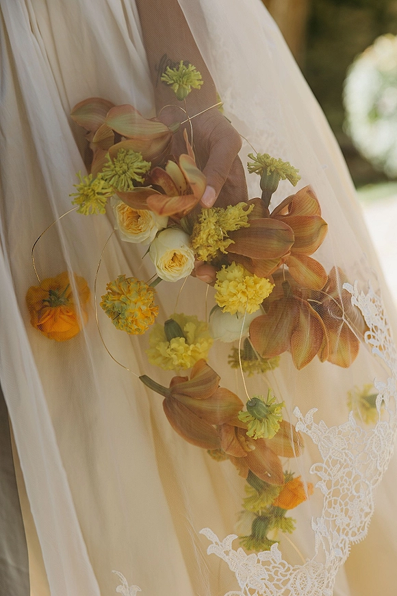 Bridal bouquet with orange and yellow bouquet blooms, roses, and ribbon loops held against lace wedding dress and veil in soft light