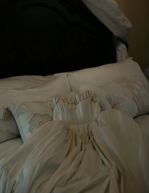 Wedding dress with a corset bodice and ruffled neckline draped in satin on a bed with decorative pillows and upholstered headboard