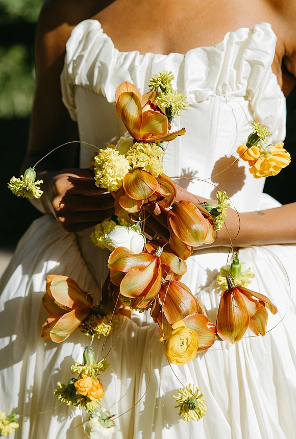 Bridal bouquet cascading with orange and yellow flowers and green stems against a white strapless wedding dress in sunlit greenery