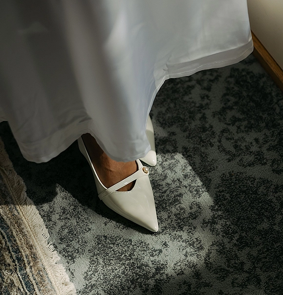 Bridal shoes, white wedding heels with pointed toes and a Mary Jane strap, gold buckle by the bridal gown hem on patterned carpet in sunlight