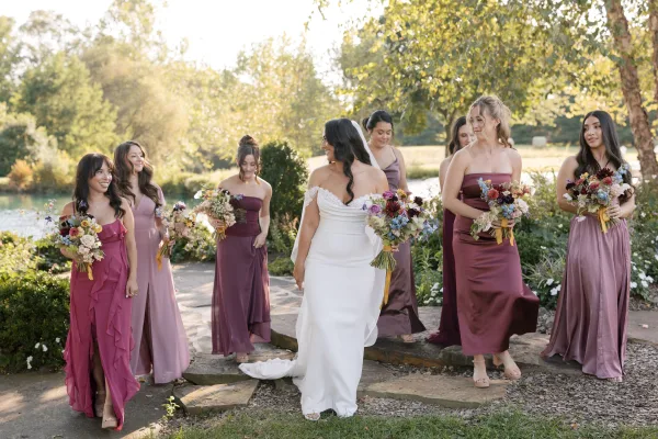 Bride and bridesmaids in mix-and-match bridesmaid dresses walk a stone path, smiling with ribbon-tied bouquets, garden trees and a pond behind them.