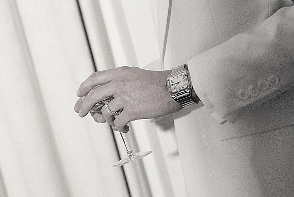 Groom detail with wedding band and wristwatch as he holds a champagne flute, suit jacket sleeve lit by soft window light indoors