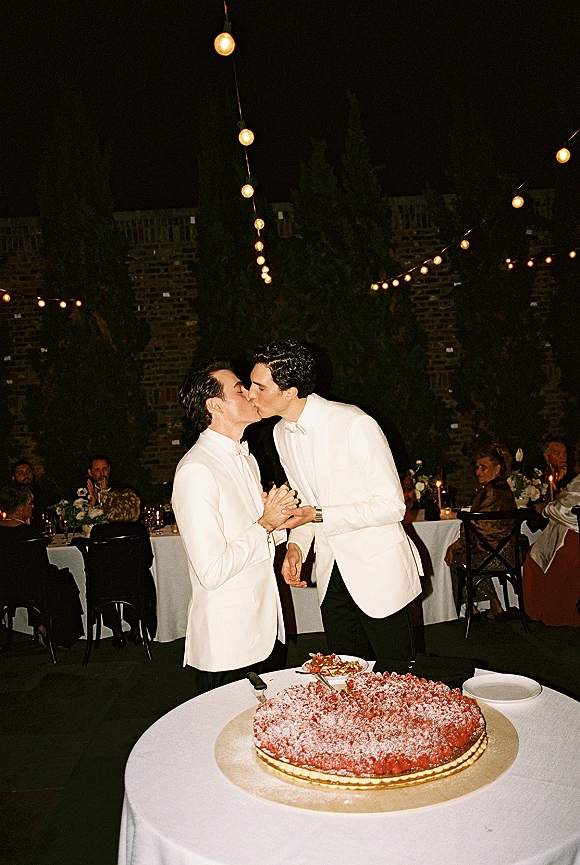 Wedding kiss between two grooms in white dinner jackets and bow ties beside a candlelit cake table under string lights at night reception