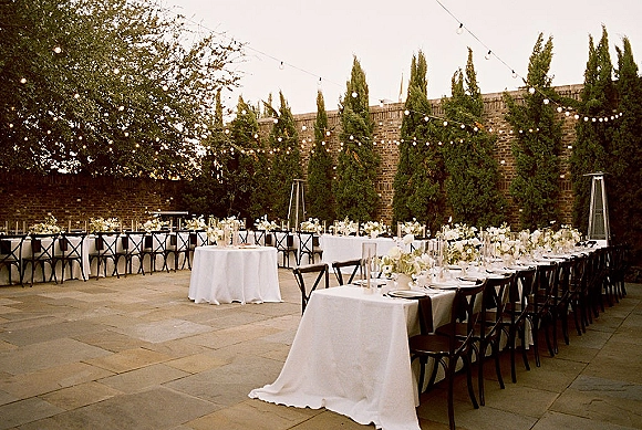 Outdoor reception setup with al fresco wedding reception style, long banquet tables under string lights by a brick wall and evergreens