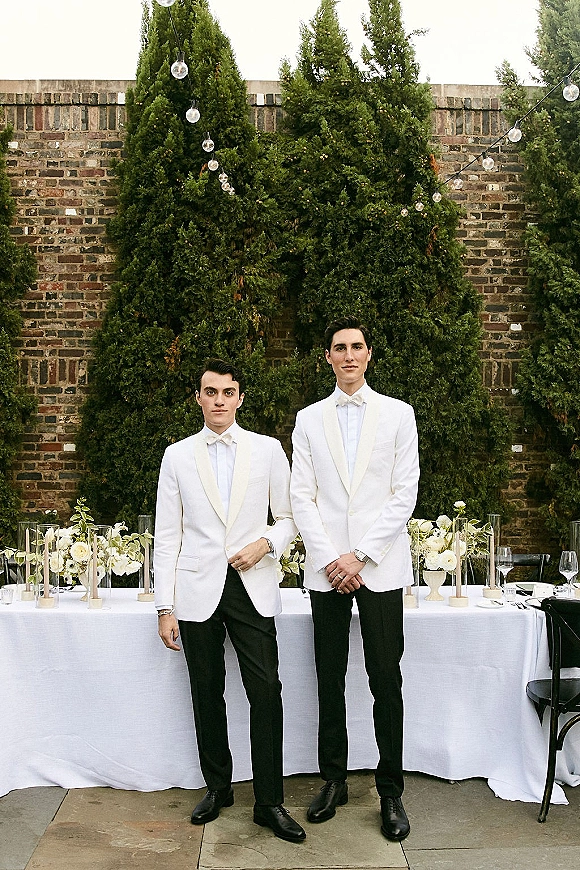 Groom portrait in a white tuxedo jacket with bow tie and boutonniere beside a candlelit reception table against a brick wall backdrop
