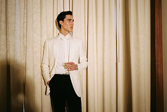 Groom portrait in a white tuxedo groom look, wearing a bow tie and holding a cocktail glass beside cream curtains in warm light