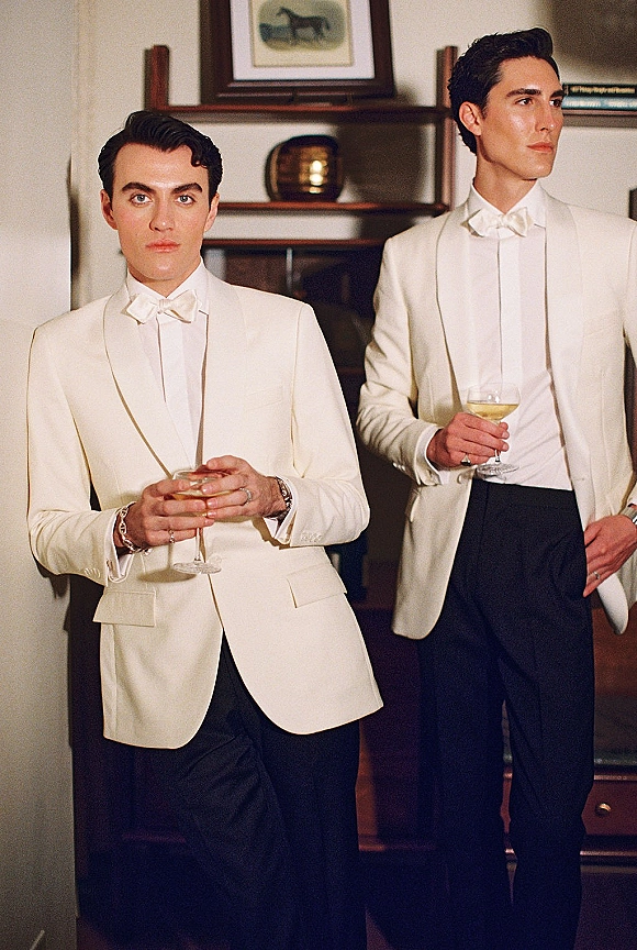 Groom portrait in a white tuxedo with bow tie and cummerbund, holding a cocktail glass by wood shelves with framed artwork indoors