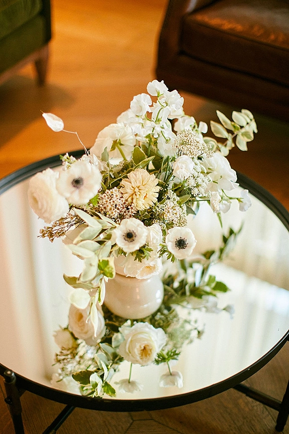Wedding centerpiece with white floral centerpiece blooms and greenery in a ceramic vase on a round coffee table in a lounge setting