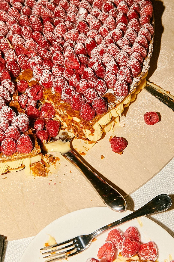 Wedding cake topped with raspberries and powdered sugar, with a slice on a dessert plate beside a cake knife on a tablecloth