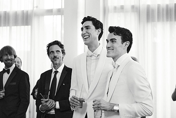 Groom portrait of two grooms in white tuxedos laughing with cocktail glasses near sheer curtains as wedding guests mingle in soft window light