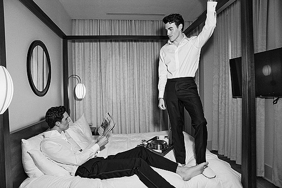 Groom getting ready in a hotel room, lounging on the bed in a dress shirt and black trousers, reading a newspaper beside room service coffee