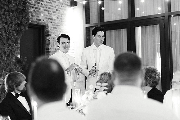 Wedding speech as two grooms in white tuxedos raise champagne flutes at the head table, brick wall and string lights behind guests