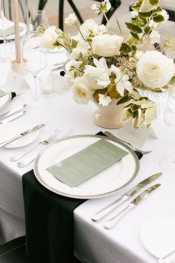 Reception tablescape with wedding table centerpiece of white flowers and greenery in a compote vase, taper candles, and black napkins on gray runner