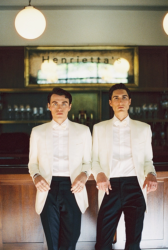 Groomsmen portrait in white dinner jackets and bow ties, standing at a bar with globe lights, glassware shelves, and a framed sign behind them