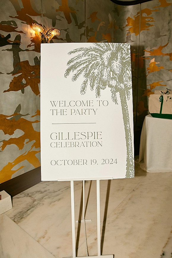 Wedding welcome sign on an easel with green serif text and palm tree illustration, set in an indoor entryway with gift bags nearby