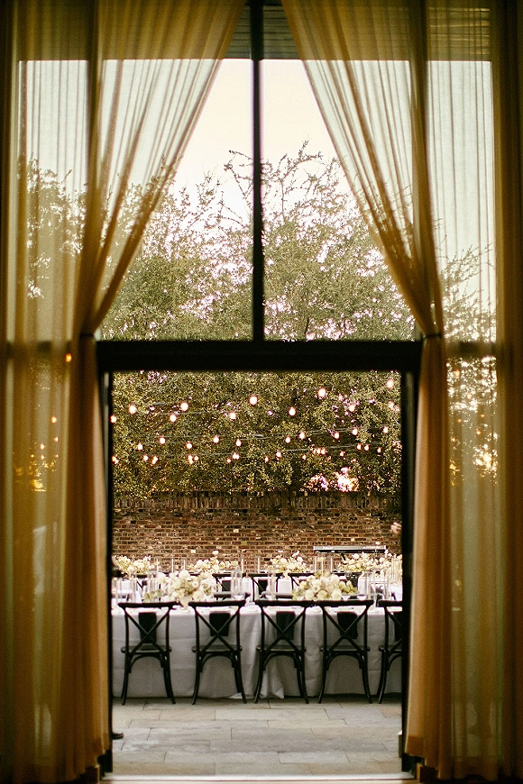 Reception tablescape on a long banquet wedding table with white linens, floral centerpieces and candles under string lights by brick wall windows