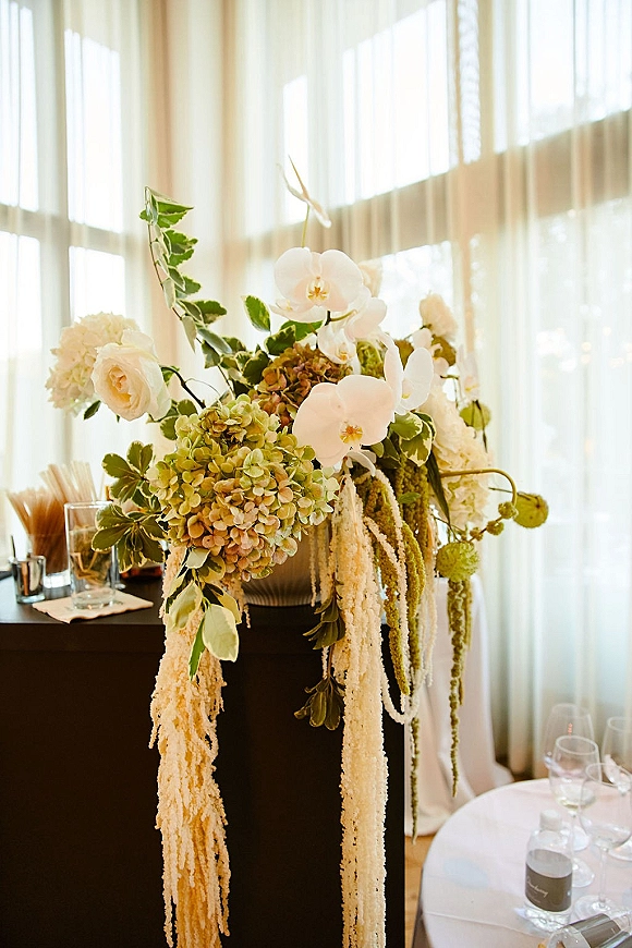 Wedding floral arrangement white orchid centerpiece with hydrangeas, roses, and hanging amaranthus in a vase on a window-lit cocktail table