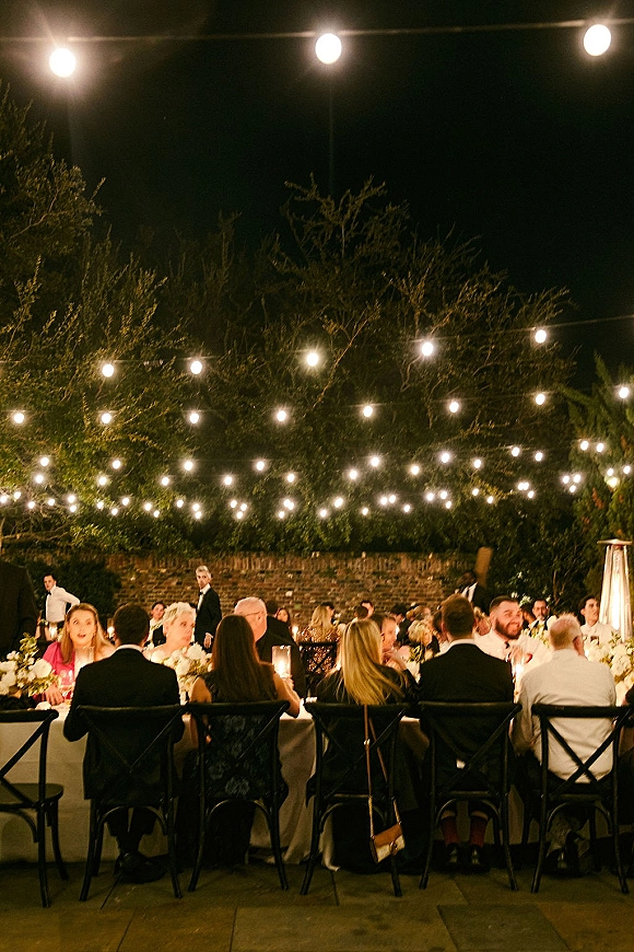 Outdoor reception dinner under string lights with candlelit long banquet table, white florals, and black crossback chairs on a patio at night