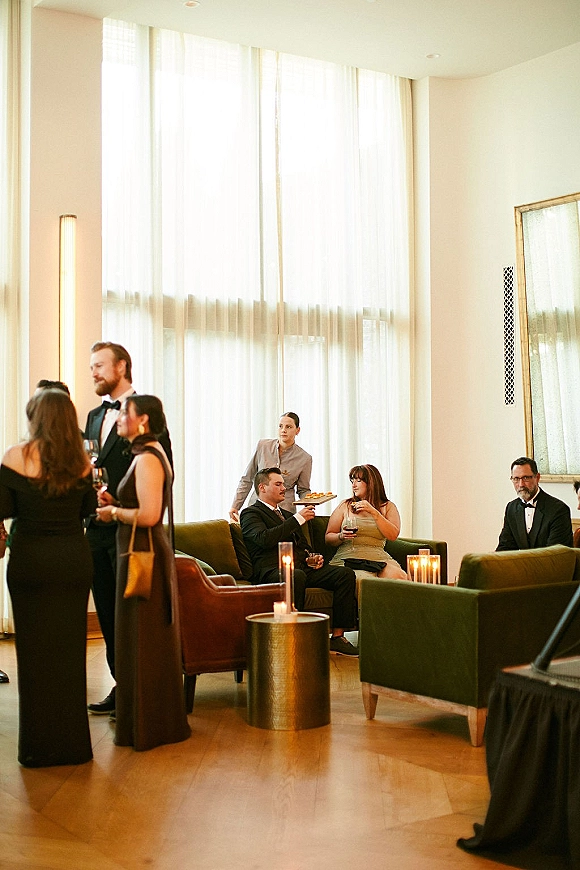 Wedding cocktail hour with guests in cocktail dresses and tuxedo holding wine glasses as a server passes canapés in a candlelit lounge by tall windows