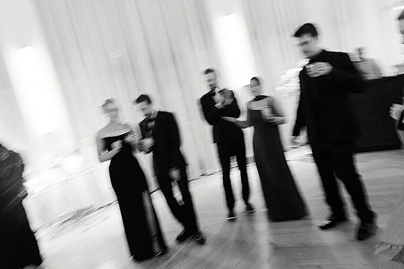 Wedding reception guests in black tie attire holding drink glasses and mingling on a dance floor with draped fabric walls and banquet lighting