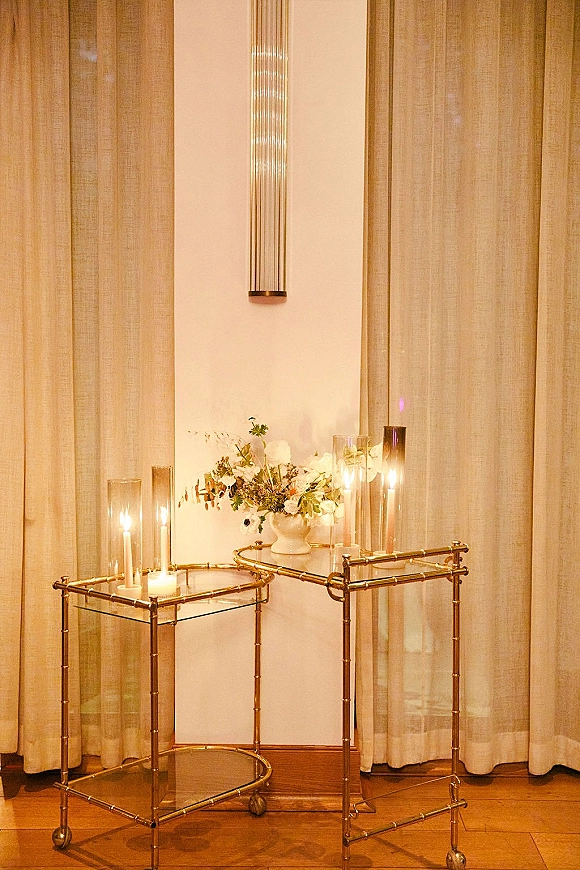 Wedding bar cart with gold bar cart decor, glass shelves, pillar candles in hurricanes, and a white floral vase by sheer curtains and sconce