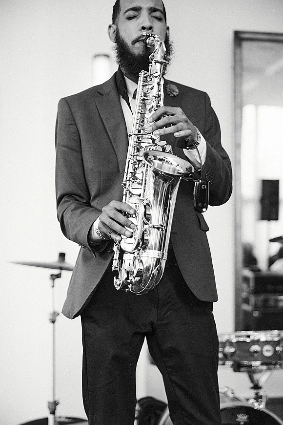 Wedding saxophonist playing live wedding music in a suit and tie with boutonniere on an indoor stage beside a drum set and mic stand