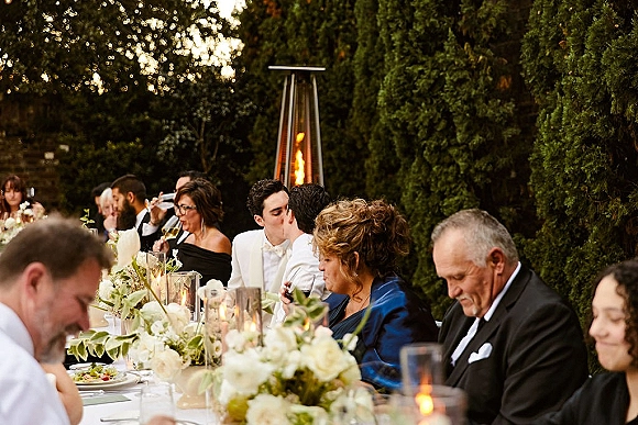 Reception dinner at a long banquet table with white tablecloth, white floral centerpieces and cylinder candles in an outdoor garden setting
