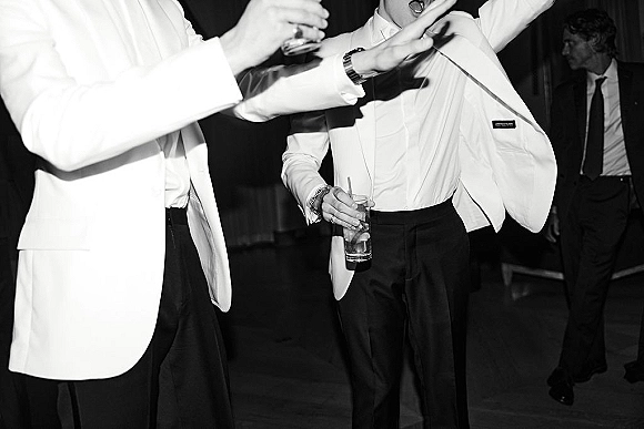 Wedding reception dancing as groomsmen dance in white tuxedo jackets and black bow ties, holding drinks on a dim reception dance floor