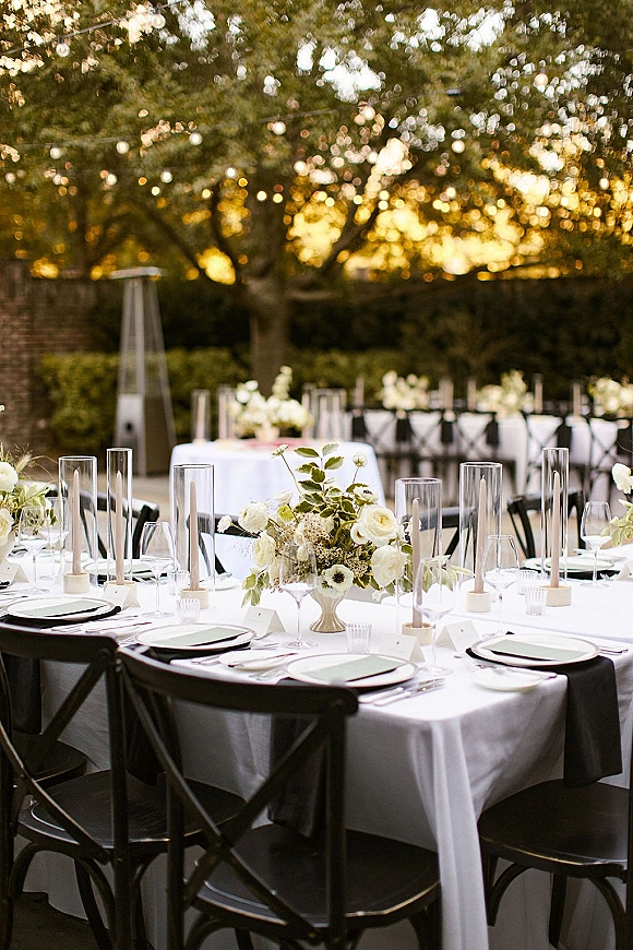 Reception tablescape with a round wedding table setup featuring a white floral centerpiece, roses and candles under string lights on a garden patio