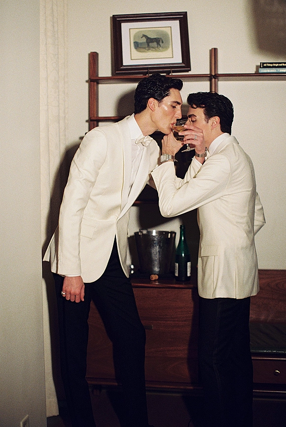 Groom portrait in a white dinner jacket and bow tie holding a champagne coupe, standing by a vintage bar cabinet with wine bottles and bookshelves