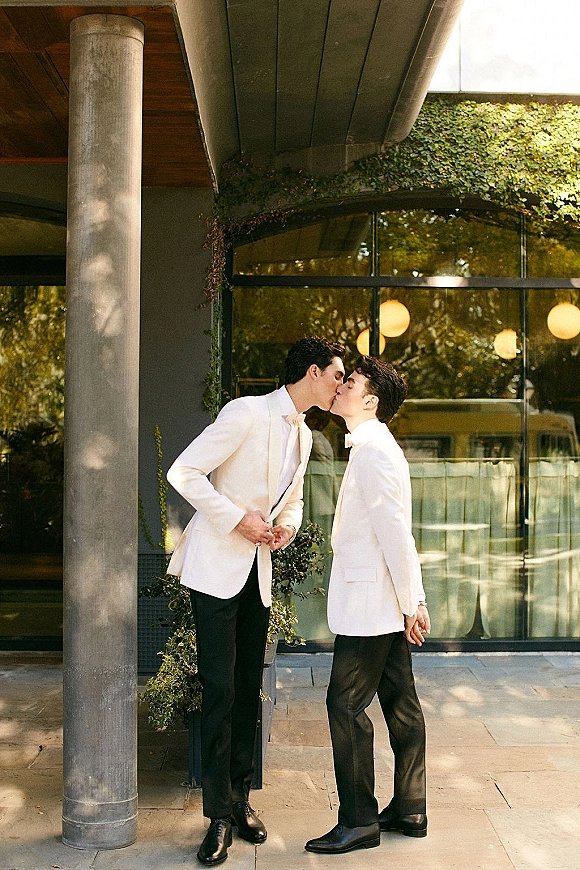 Wedding kiss portrait of two grooms in white dinner jackets and bow ties kissing by a concrete column with ivy wall and glass windows
