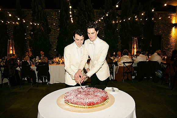 Wedding cake cutting by two grooms in white dinner jackets with bow ties, slicing at a round table under string lights in a brick courtyard at night