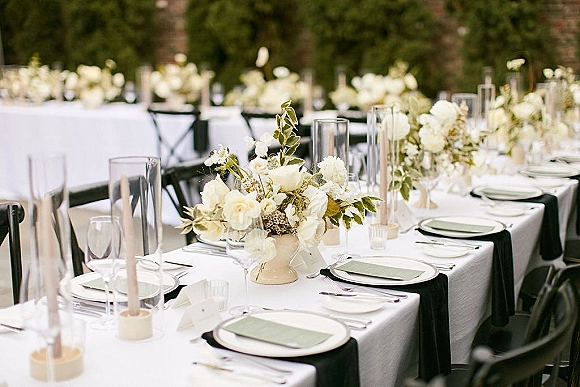 Reception tablescape with long banquet table setup, white floral centerpieces, taper candles, and black runner on an outdoor patio with hedges