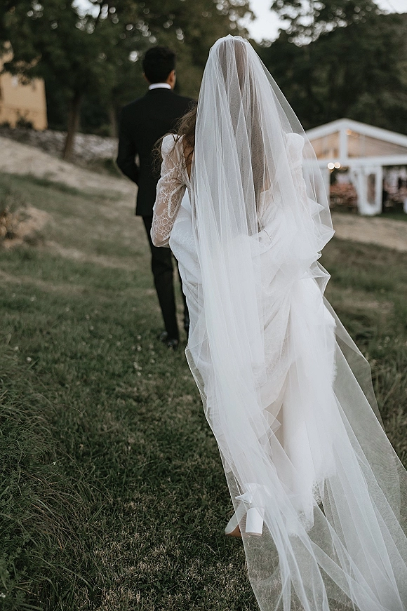First look moment as bride in lace-sleeve wedding dress and long veil walks up behind groom in black tux on a lawn near tents