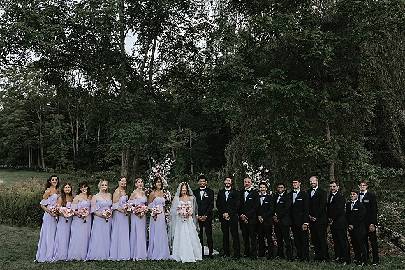 Wedding party portrait of bride with bridesmaids and groomsmen, lavender dresses and black tuxedos, under a floral arch on a garden lawn