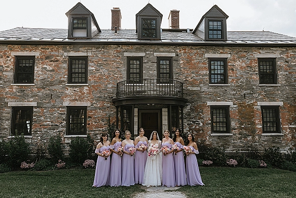 Bridesmaids group photo with the bride, featuring lavender bridesmaid dresses and pink bouquets in front of a stone estate facade