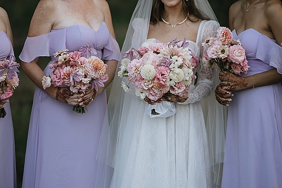 Bridesmaids and bride in lavender bridesmaid dresses hold pink and white bouquets, showing henna hands, lace gown, veil, and pearls outdoors