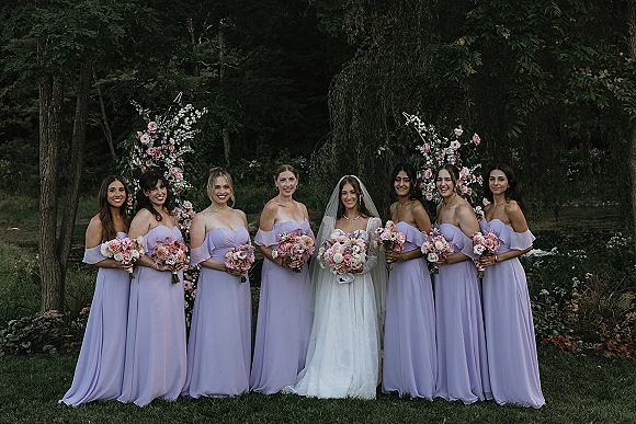 Bride with bridesmaids in lavender bridesmaid dresses holding bouquets beneath a floral arch on a green garden lawn outdoors
