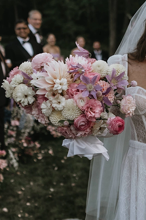Bridal bouquet of pink and lavender bouquet flowers with dahlia and ranunculus, wrapped in ribbon, held against a lace dress outdoors