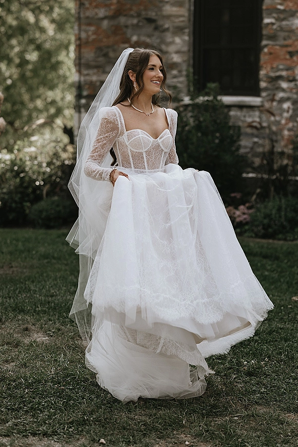Bridal portrait of a bride in a lace corset wedding dress with cathedral veil and pearls, holding her tulle skirt by a stone building in a garden