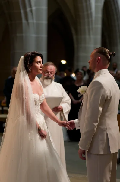 Wedding vows as the bride and groom holding hands stand in a church interior framed by stone arches and pews, her long veil trailing softly.